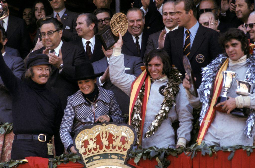 The podium (L to R): Colin Chapman (GBR) Lotus Team Owner; Marie-Helena Fittipaldi (BRA); Emerson Fittipaldi (BRA) Lotus winner; Francois Cevert (FRA) Tyrrell second. Spanish dignitaries in the back include Juan Antonio Samaranch, the provincial political head of Barcelona and member of the International Olympic Committee (IOC) and HRH King Juan Carlos of Spain. Spanish Grand Prix, Montjuich Park, 29 April 1973. From Lotus 72 by Pete Lyons, published by Evro.